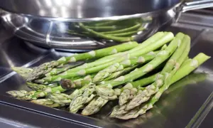 Un bouquet d'asperges vertes fraîches, prêtes pour la cuisson, repose sur une surface métallique devant une poêle.