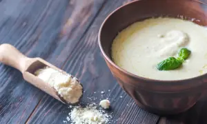 Une cuillère en bois contient de la poudre de parmesan, à côté d'un bol de crème de parmesan recette, garnie de basilic.
