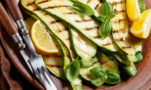 Tranches de courgette plancha grillées, garnies de basilic frais et de quartiers de citron, servies sur une assiette en terre cuite avec des couverts.