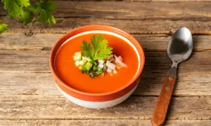 Un bol de gaspacho rouge vif garni de dés de légumes et de coriandre, prêt à être dégusté. Une cuillère repose à côté, invitant à découvrir cette recette rafraîchissante.