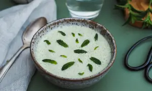 Un bol de gaspacho concombre menthe, rafraîchissant et garni de feuilles de menthe fraîche. Une cuillère et un verre d'eau sont à proximité.