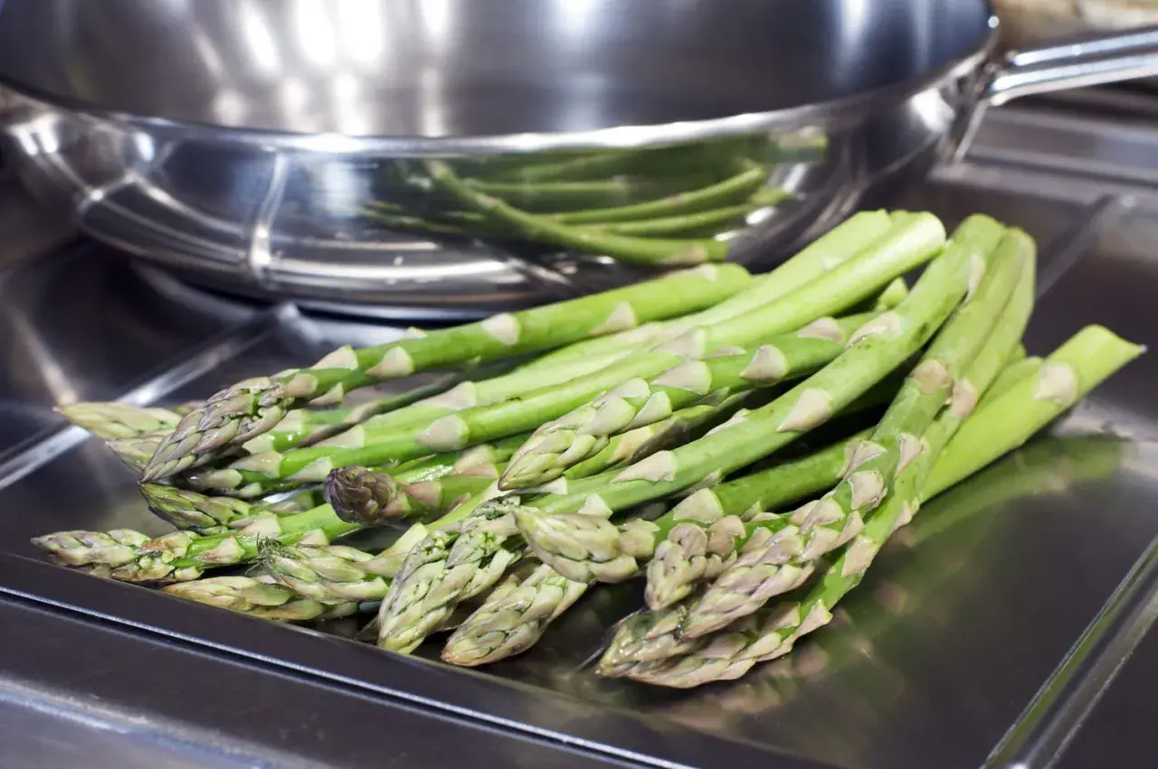 Un bouquet d'asperges vertes fraîches, prêtes pour la cuisson, repose sur une surface métallique devant une poêle.