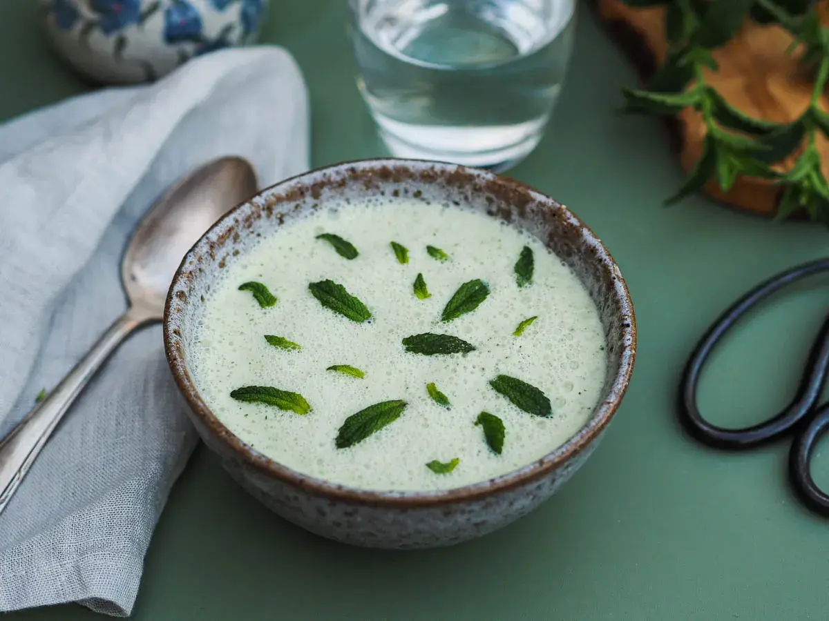 Un bol de gaspacho concombre menthe, rafraîchissant et garni de feuilles de menthe fraîche. Une cuillère et un verre d'eau sont à proximité.