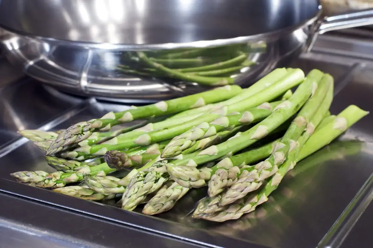 Un bouquet d'asperges vertes fraîches, prêtes pour la cuisson, repose sur une surface métallique devant une poêle.