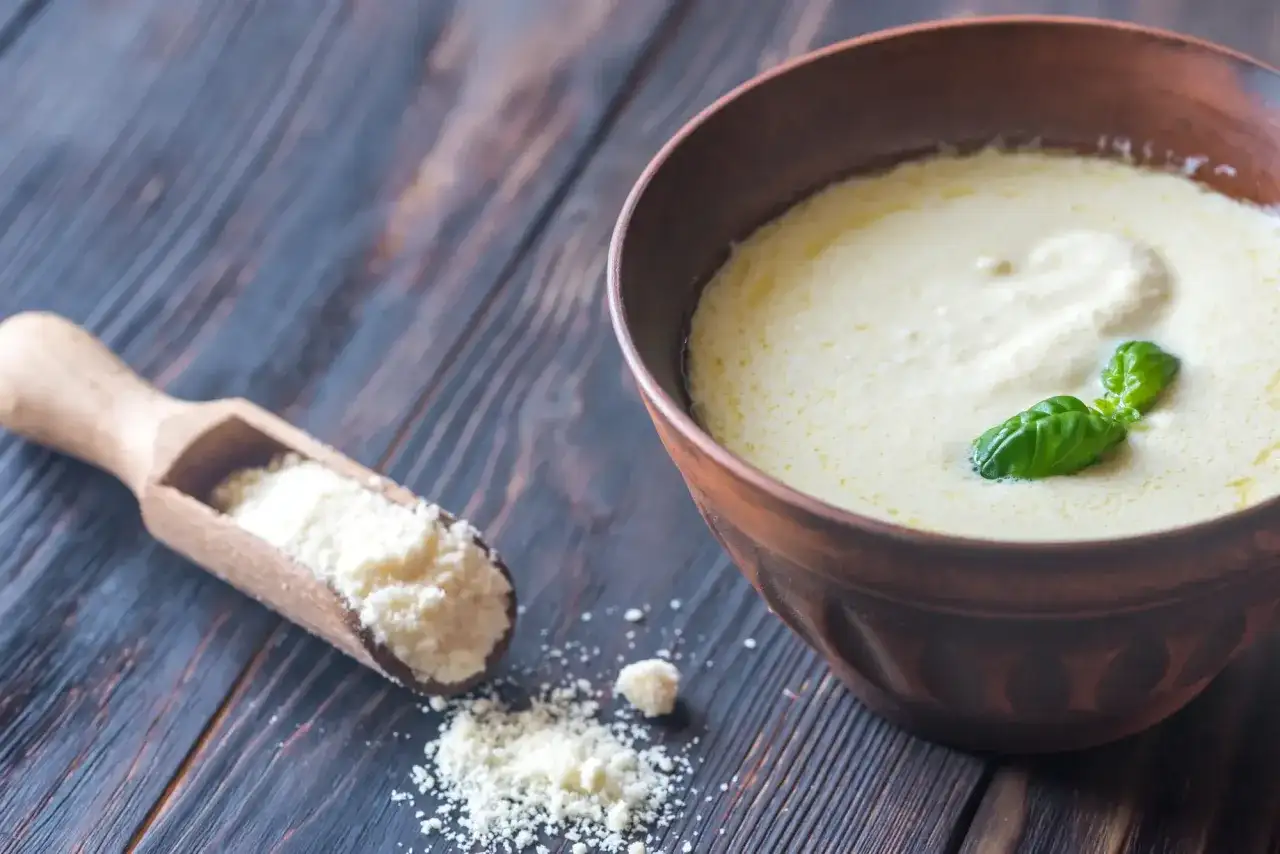 Une cuillère en bois contient de la poudre de parmesan, à côté d'un bol de crème de parmesan recette, garnie de basilic.