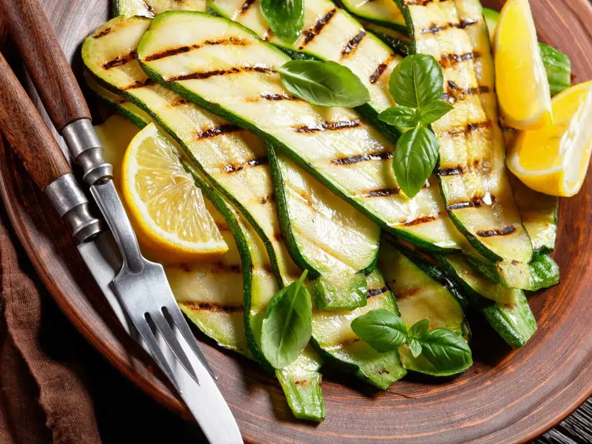 Tranches de courgette plancha grillées, garnies de basilic frais et de quartiers de citron, servies sur une assiette en terre cuite avec des couverts.