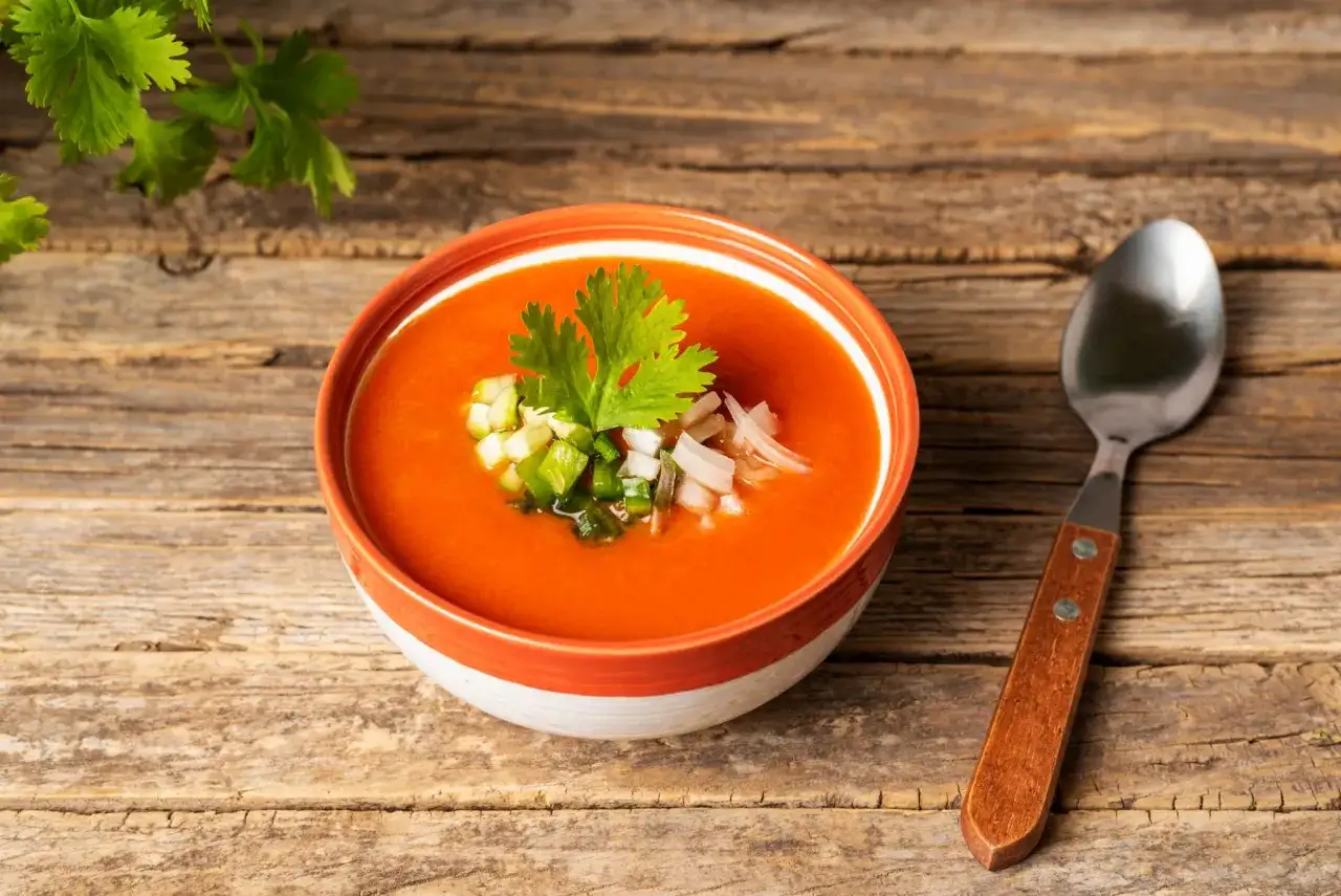 Un bol de gaspacho rouge vif garni de dés de légumes et de coriandre, prêt à être dégusté. Une cuillère repose à côté, invitant à découvrir cette recette rafraîchissante.