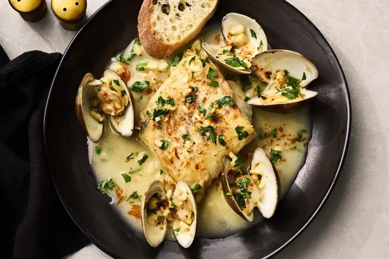 Poisson poêlé servi avec des palourdes ouvertes, baignant dans une sauce crémeuse, garni de persil et d'une tranche de pain. La cuisson des palourdes est parfaite.