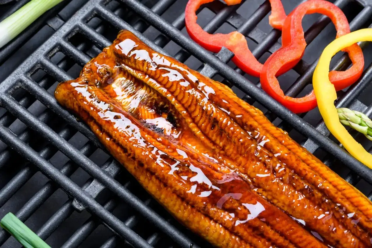 Une anguille au barbecue, laquée et brillante, est posée sur une grille avec des poivrons colorés et des asperges.