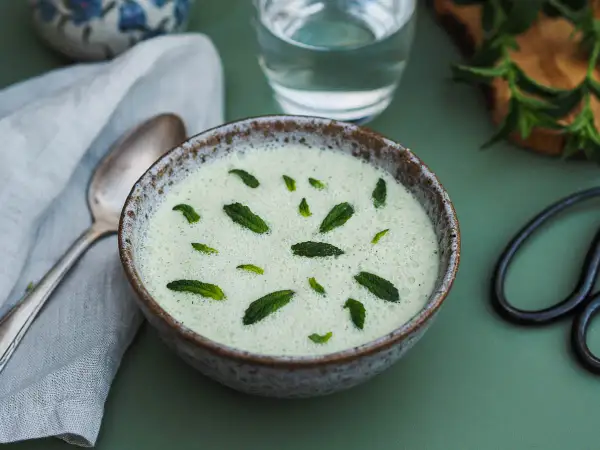 Un bol de gaspacho concombre menthe, rafraîchissant et garni de feuilles de menthe fraîche. Une cuillère et un verre d'eau sont à proximité.